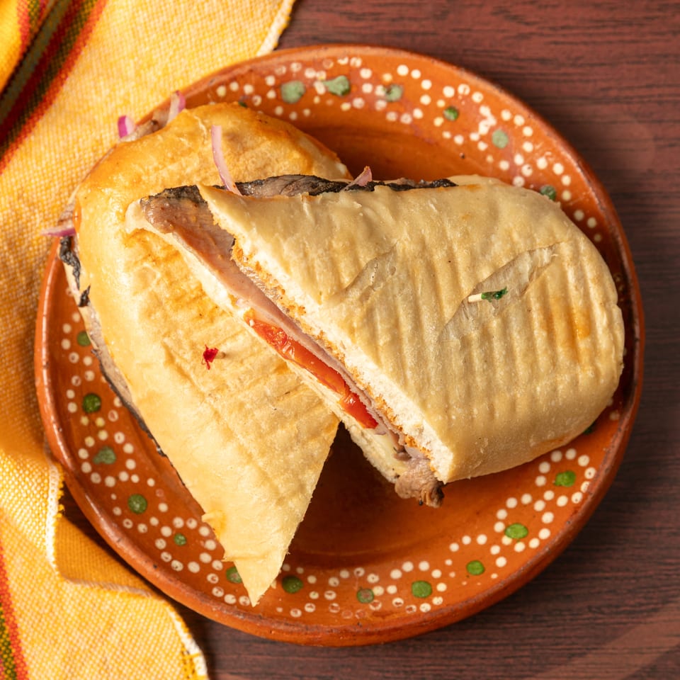 Grilled meat sandwich with tomatoes and onions on a decorative ceramic plate beside a colorful woven napkin.