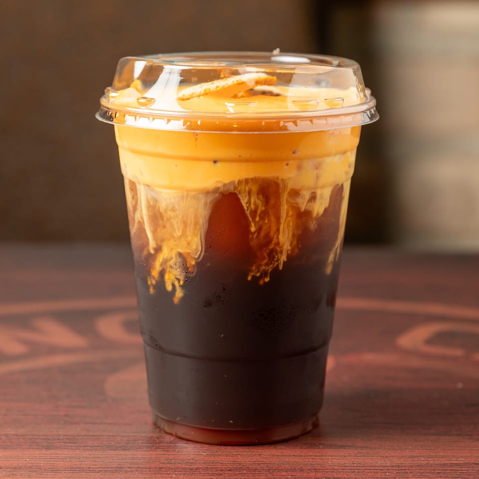 Layered iced coffee with creamy orange topping in a clear plastic cup on a wooden table.