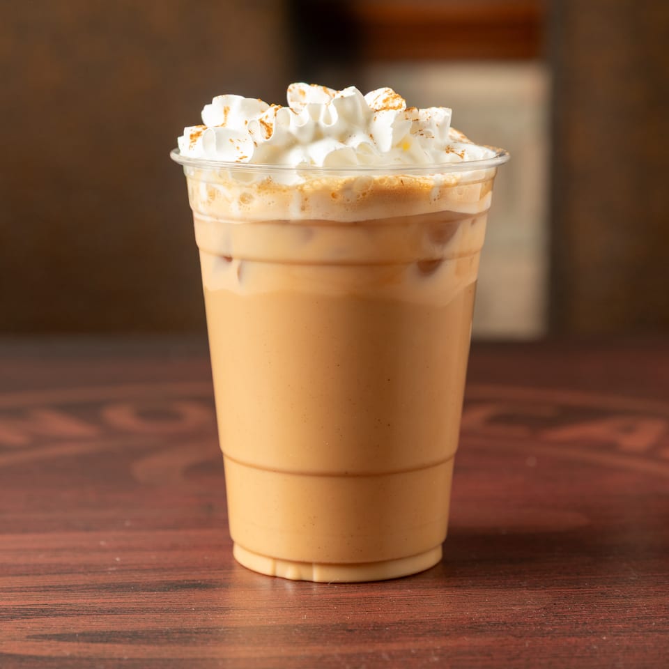 Iced coffee topped with whipped cream and cinnamon sits in a clear cup on a wooden table.