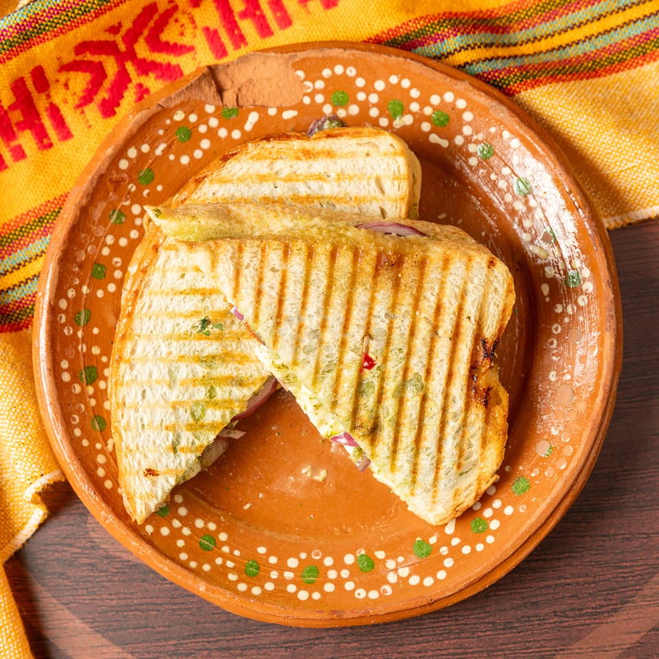 Grilled panini sandwich with visible vegetables on a decorative terracotta plate beside a colorful woven cloth.