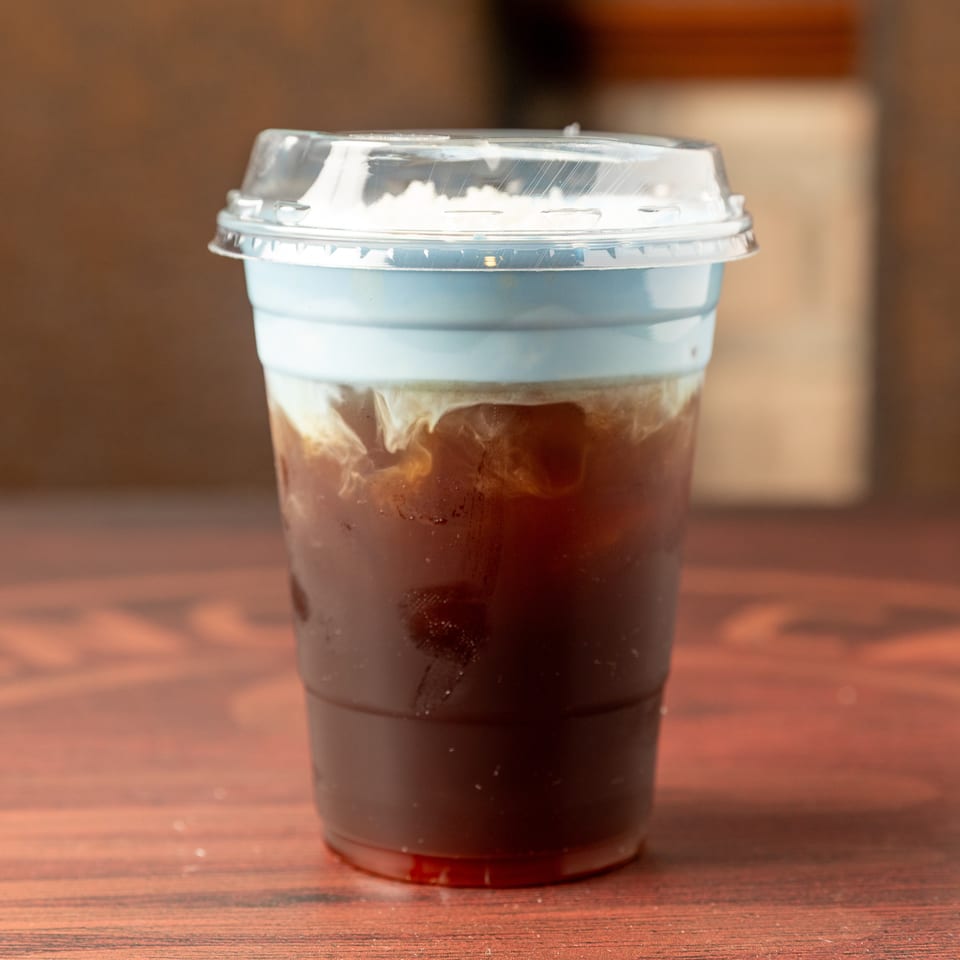 Iced dark coffee in a clear plastic cup with a domed lid, resting on a warmly lit wooden table.