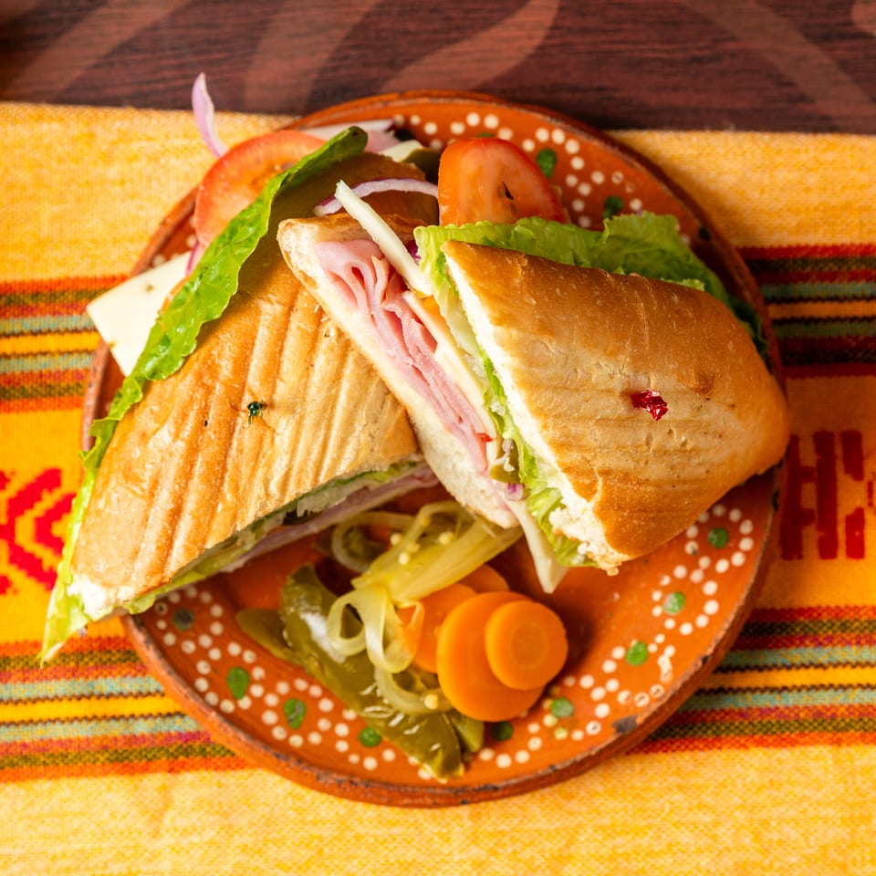 Ham and cheese sandwiches with lettuce, tomato, onion, pickles, and carrots on a colorful patterned plate.