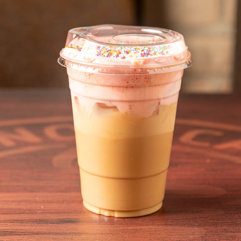 Layered creamy orange drink in a clear plastic cup, topped with whipped cream and colorful sprinkles on a wooden table.