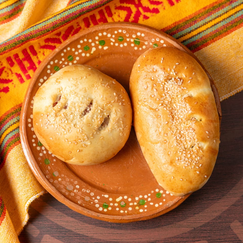 Two sesame-topped golden breads sit on a decorated earthenware plate beside a colorful striped cloth.